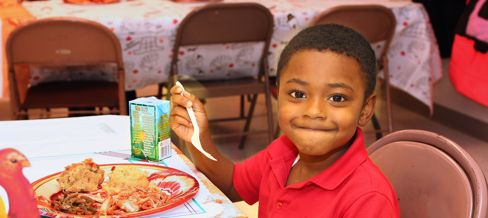 A smiling child is holding a fork over a plate of food with a juice box on the table.