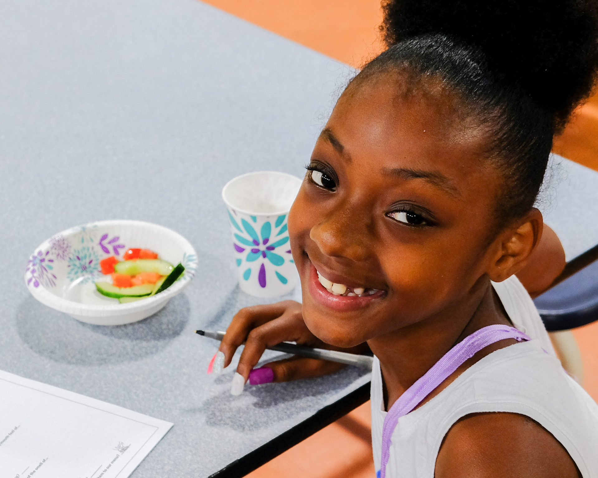 A smiling individual is seated at a table with a bowl of salad, a cup, and papers, holding a writing instrument.