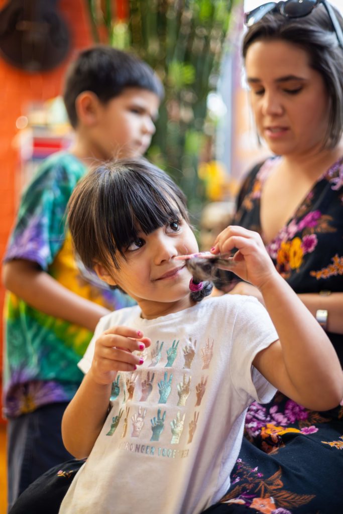 A child looks curiously at the camera while holding a paintbrush, with focused individuals in the background.