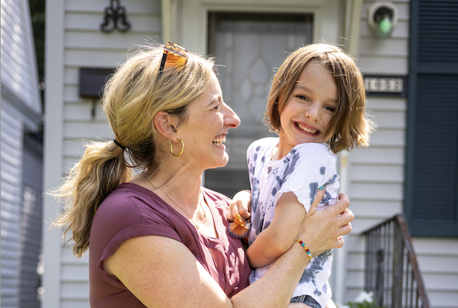 A smiling adult is holding a happy child in front of a house with a porch.