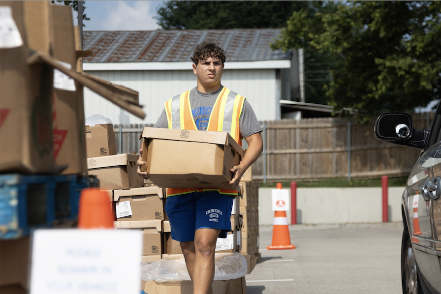 A person wearing a yellow reflective vest is carrying a cardboard box outdoors with a background of piled boxes and a building.
