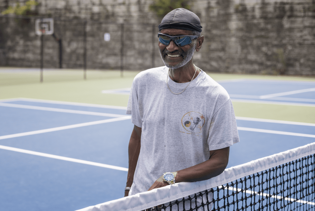 A smiling person is standing by a tennis net wearing sportswear and sunglasses on a sunny day.