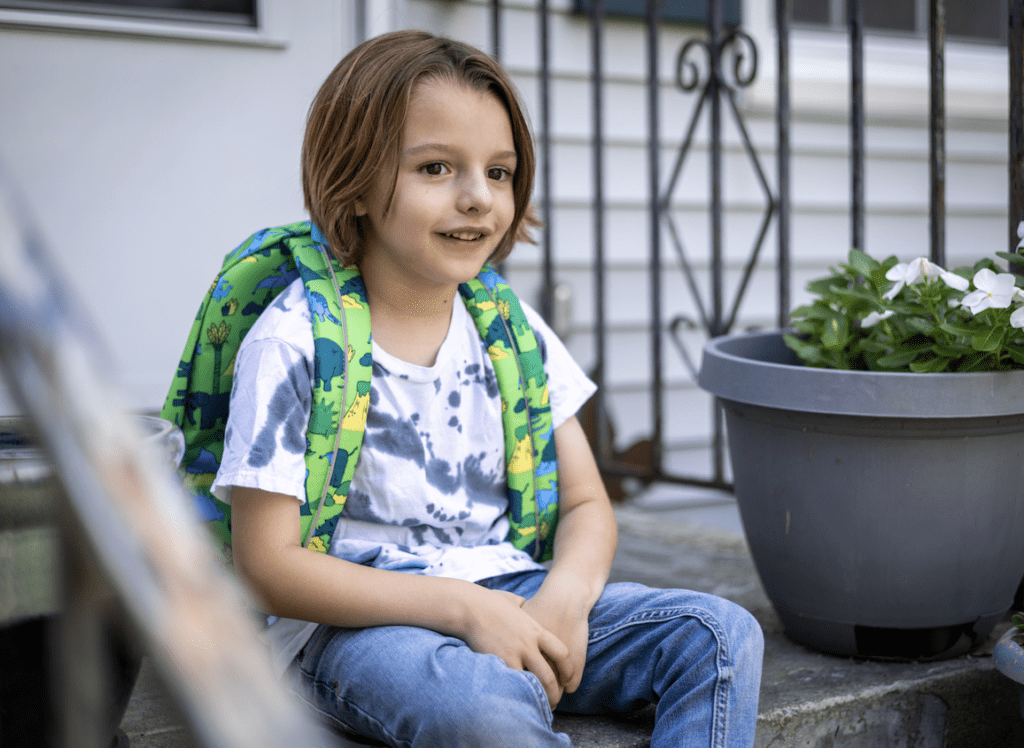 A child sits on a step outdoors wearing a backpack and a casual outfit with a subtle smile on their face.