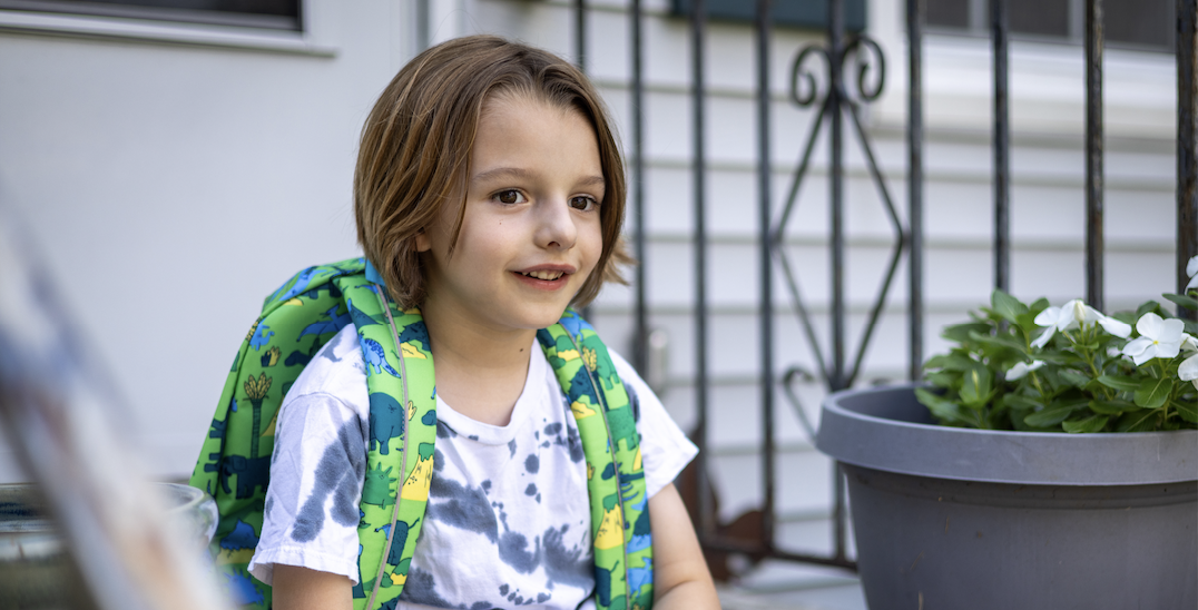 A smiling child with a backpack is sitting on steps outside a building.