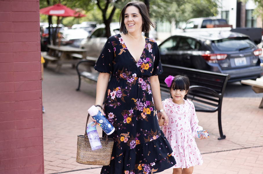 A smiling individual in a floral dress walks hand in hand with a child wearing a pink dress on a city street.
