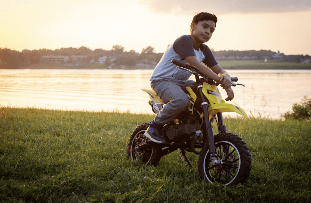 A person on a small motorcycle poses on a grassy field with a body of water at dusk in the background.