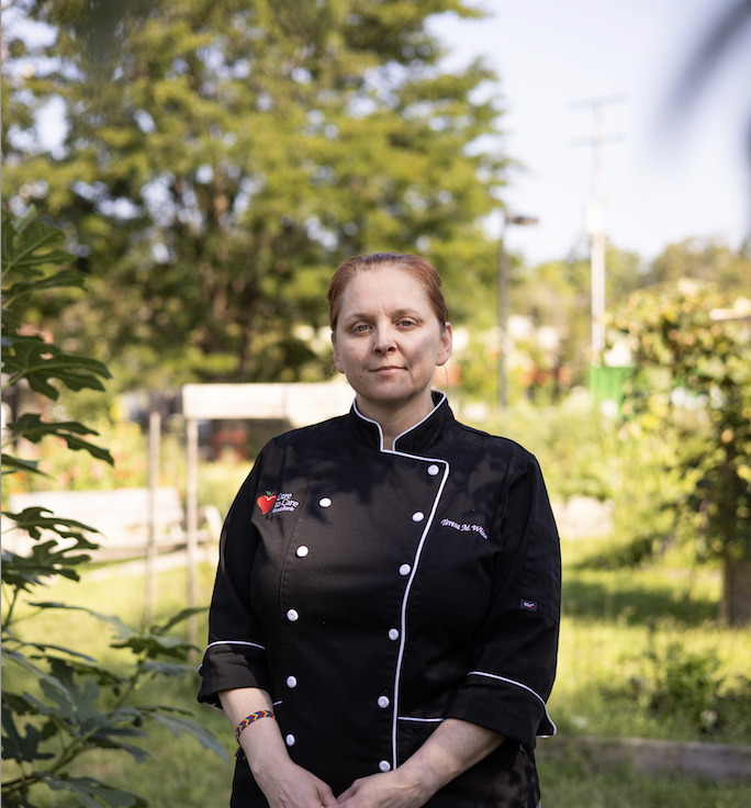 A professional chef stands outdoors dressed in a black chef's coat, smiling at the camera.