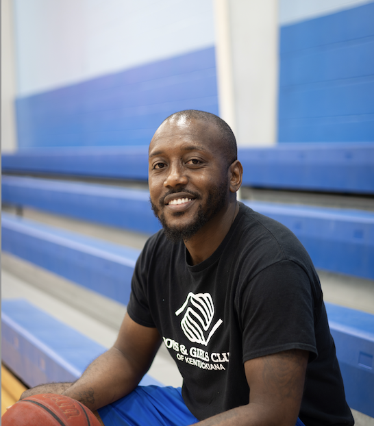 A smiling individual is seated in a gymnasium with blue bleachers in the background.
