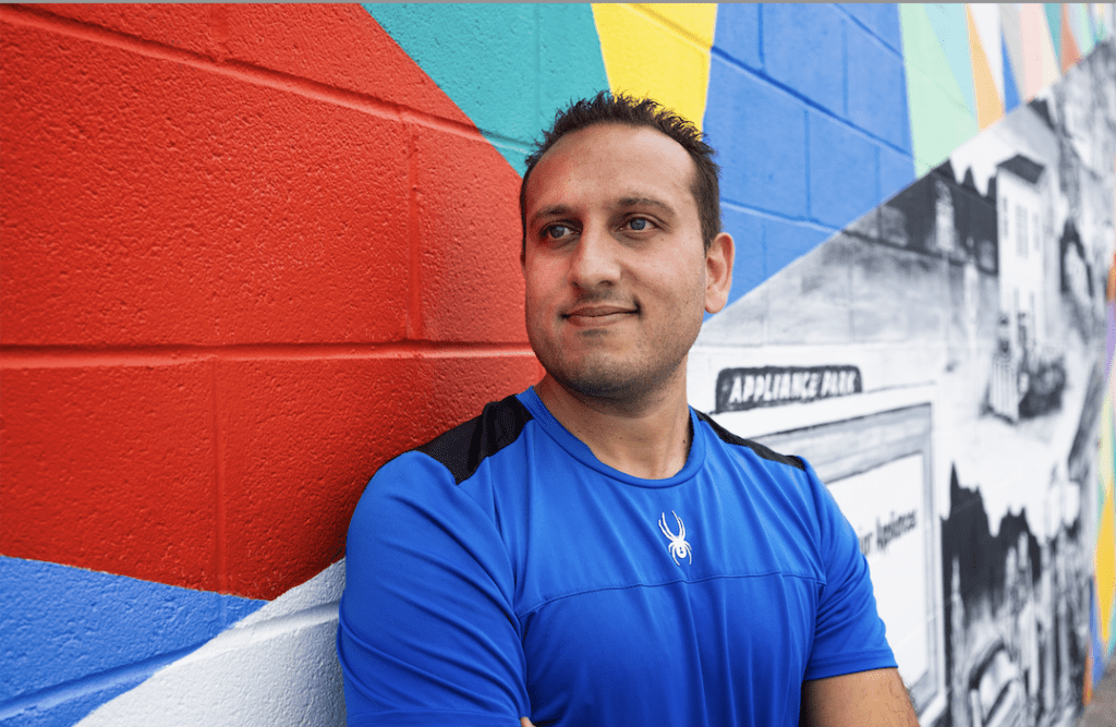 A smiling individual wearing a blue top leans against a vibrantly colored geometric wall mural.
