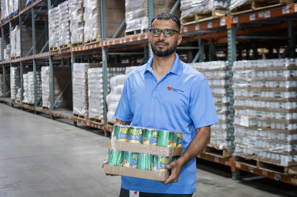 A person wearing a blue polo shirt with a logo on it stands holding a cardboard box in a warehouse with palletized goods in the background.