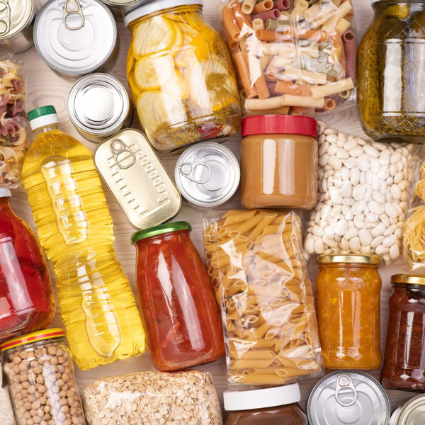 A variety of preserved and packaged food items, including bottles, cans, jars, and bags, are laid out showcasing a mix of condiments, grains, and snacks.