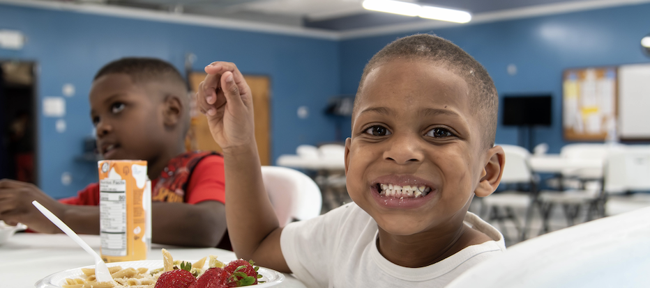 A smiling child is holding up a grape with a plate of food in front of them in a room with blue walls.