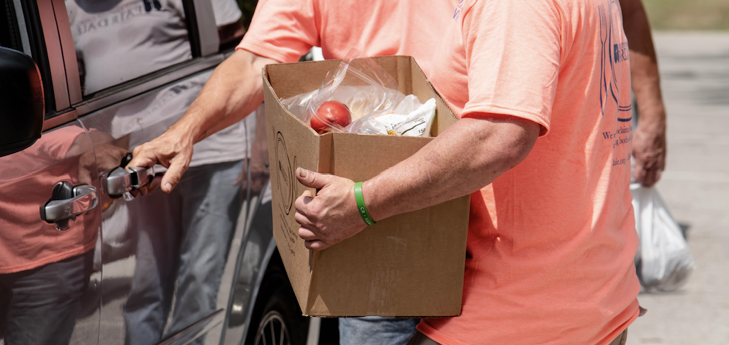 A person in a pink shirt is holding a cardboard box filled with groceries, highlighting a charitable activity or food donation.