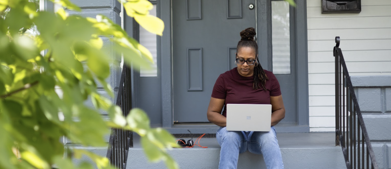 A person is sitting on a doorstep working on a laptop with green foliage in the foreground.