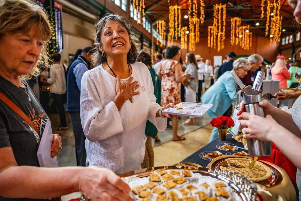 A joyful person is offering a tray of appetizers at an indoor event with hanging decorations and a crowd in the background.