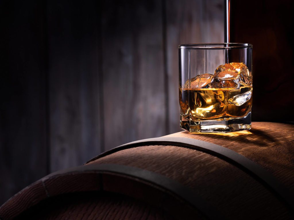 A glass of whiskey with ice sits atop a curved wooden surface against a dark backdrop.
